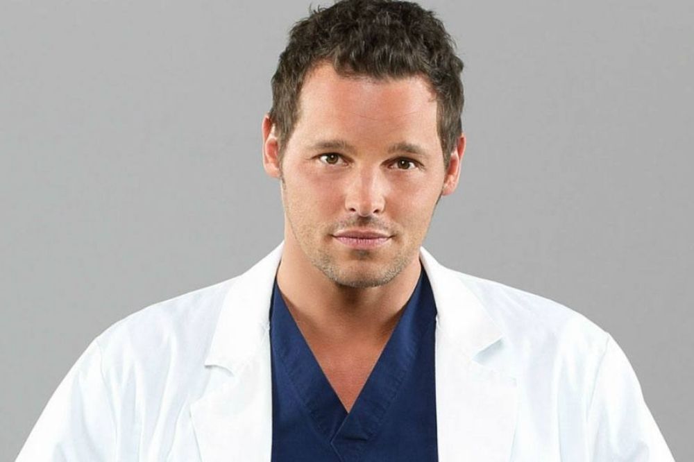 Foto de Justin Chambers como o médico Alex Karev, seu personagem em Grey's Anatomy; ele usa um jaleco branco e uniforme azul