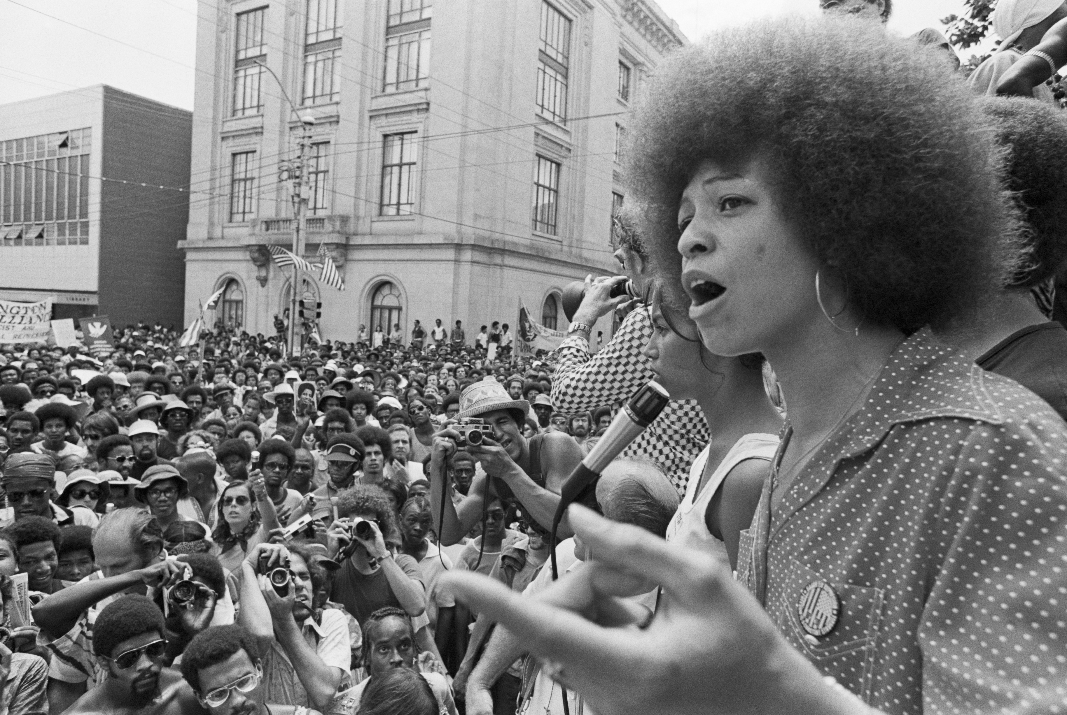 Angela Davis, à direita, discursando para um público em 1974. A foto é em preto e branco.