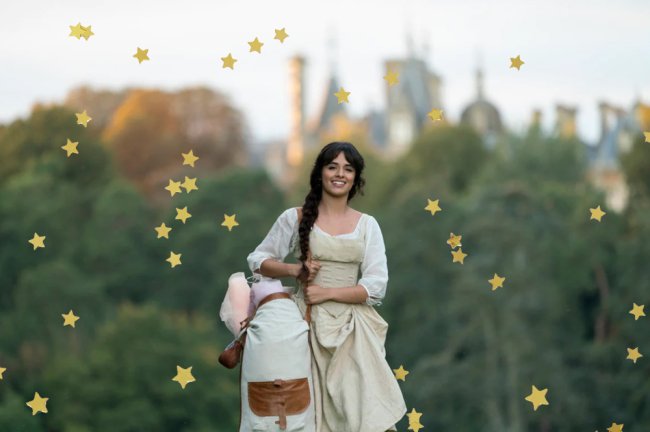 Camila Cabello, no papel de Cinderella, caminhando por uma gramado, segurando um saco de tecidos e com um sorisso no rosto. Atrás dela, aparece o castelo do Príncipe