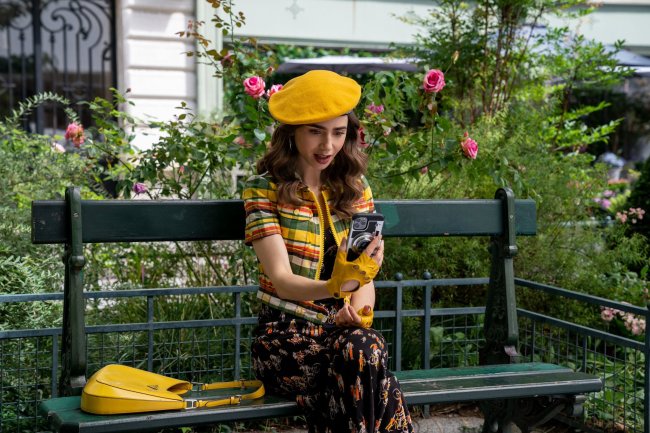 Lily Collins como Emily em imagem de da segunda temporada de Emily em Paris; ela está sentada em um banco na rua com paisagem verde com flores rosas ao fundo; ela também está olhando para o celular com expressão chocada e usa um look com detalhes amarelos como boina, luva e a bolsa colocada ao seu lado no banco
