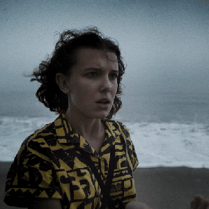 Gif de Millie Bobby Brown como Eleven na série Stranger Things. Ela aparece com blusa estampada amarela e preta em uma praia.