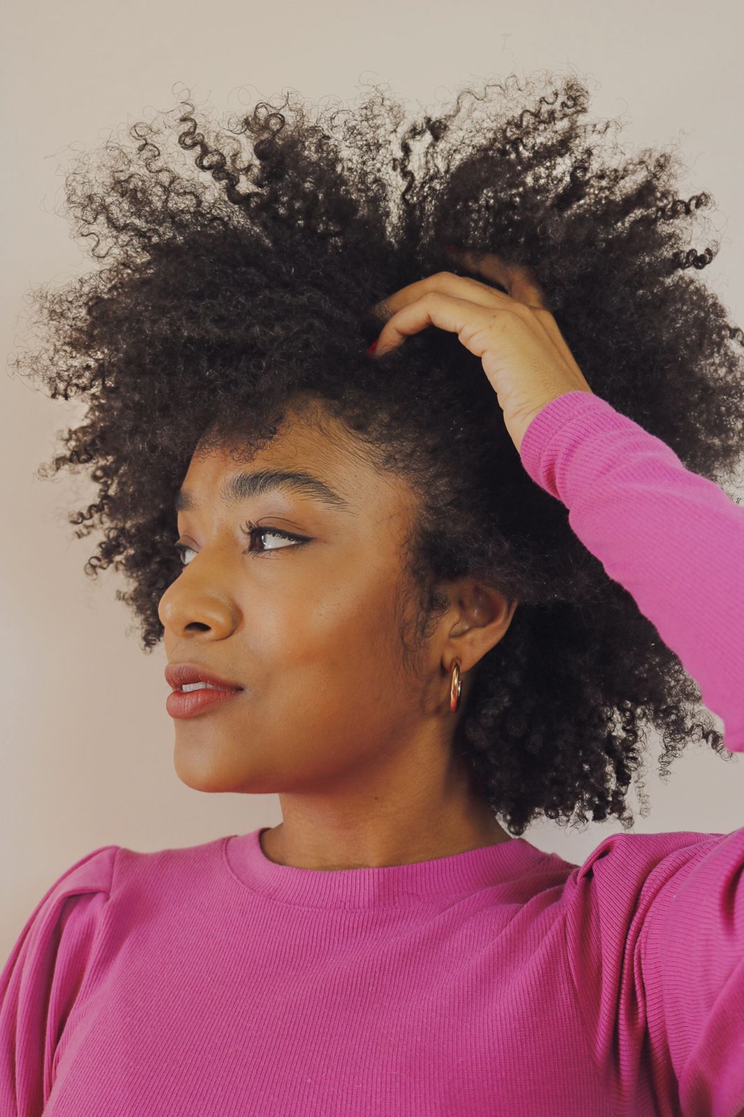 sah-oliveira-cabelo-crespo A influenciadora Sah Oliveira usando blusa rosa de manga longa, olhando para o lado com uma das mãos em seu cabelo crespo