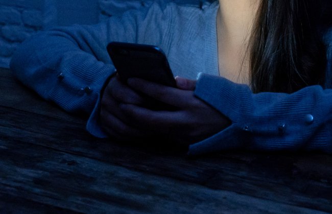 Foto de uma menina com os braços apoiado em uma mesa, segurando um celular. Não dá para ver o rosto dela e a imagem está escura