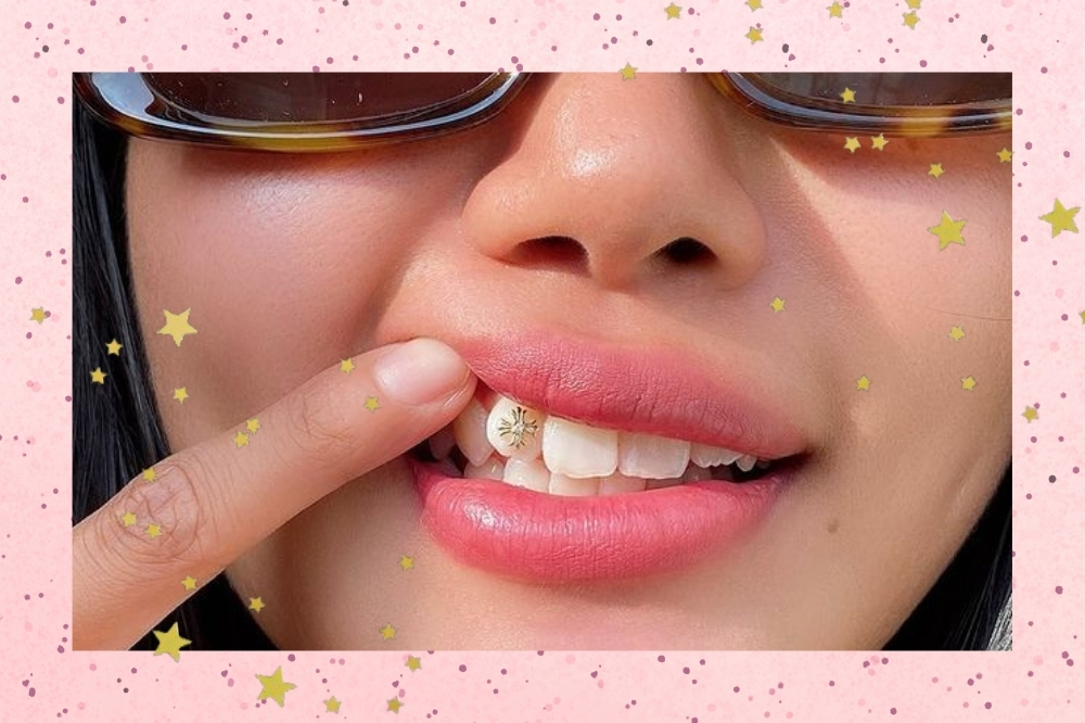 Montagem com o fundo rosa com bolinhas e detalhe de estrelas douradas com a foto de uma mulher. A foto é de um close no rosto de uma mulher mostrando um piercing no dente dela, ela usa o dente indicador para levantar os lábios para mostrar o piercing e usa um óculos de sol retangular.
