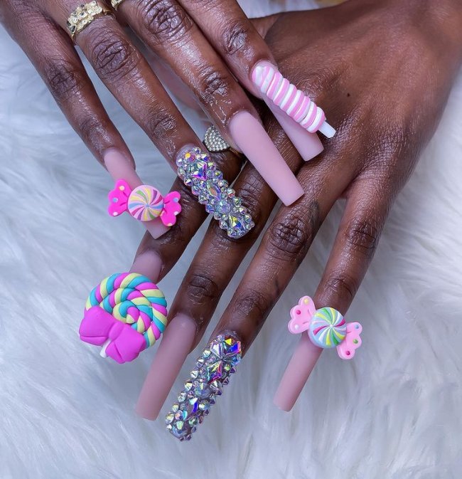 unhas-decoradas-doces Foto de unhas decoradas. Todos as unhas estão com esmalte rosa claro e detalhes 3D de docinhos coloridos, o dedo anelar está decorados com cristais.