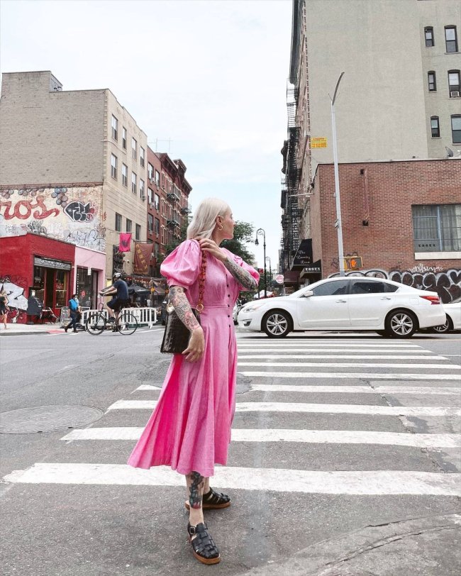 Jovem de lado posando em faixa para pedestre, ela usa um vestido longo cor de rosa e uma bolsa preta.