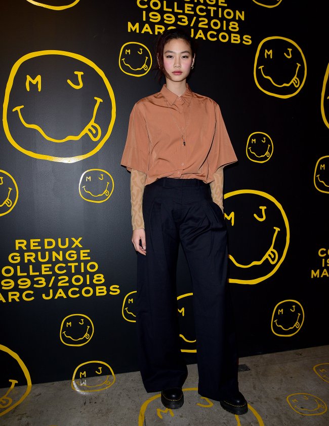 Jung Ho-yeon posando para foto em frente a mural preto com slime em amarelo. Sua expressão é séria e ela uma das mãos no bolso.