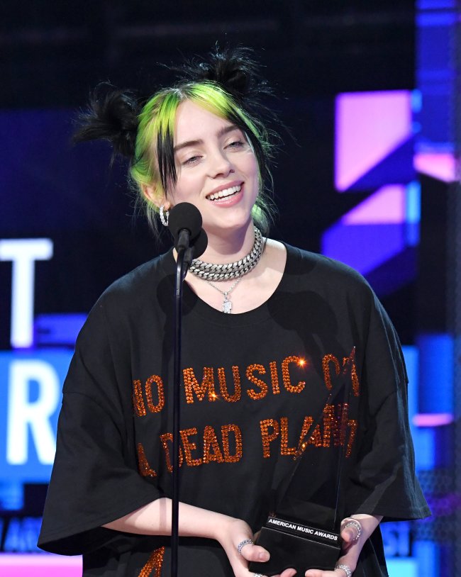 Billie Eilish segurando prêmio com camisa preta e expressão feliz.