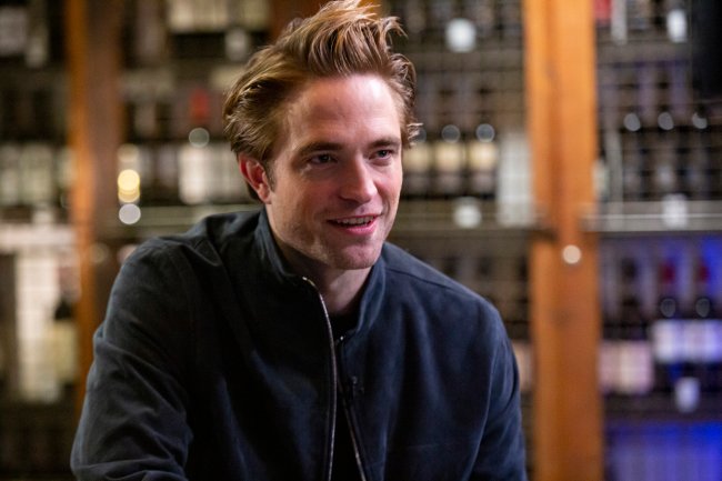 Foto de Robert Pattinson olhando na direção da direita e sorrindo. Ele usa uma jaqueta azul escura e o fundo está desfocado.