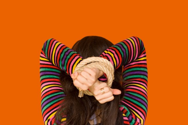 Foto de uma menina adolescentes de costas, com as mãos amarradas por uma corda atrás da cabeça