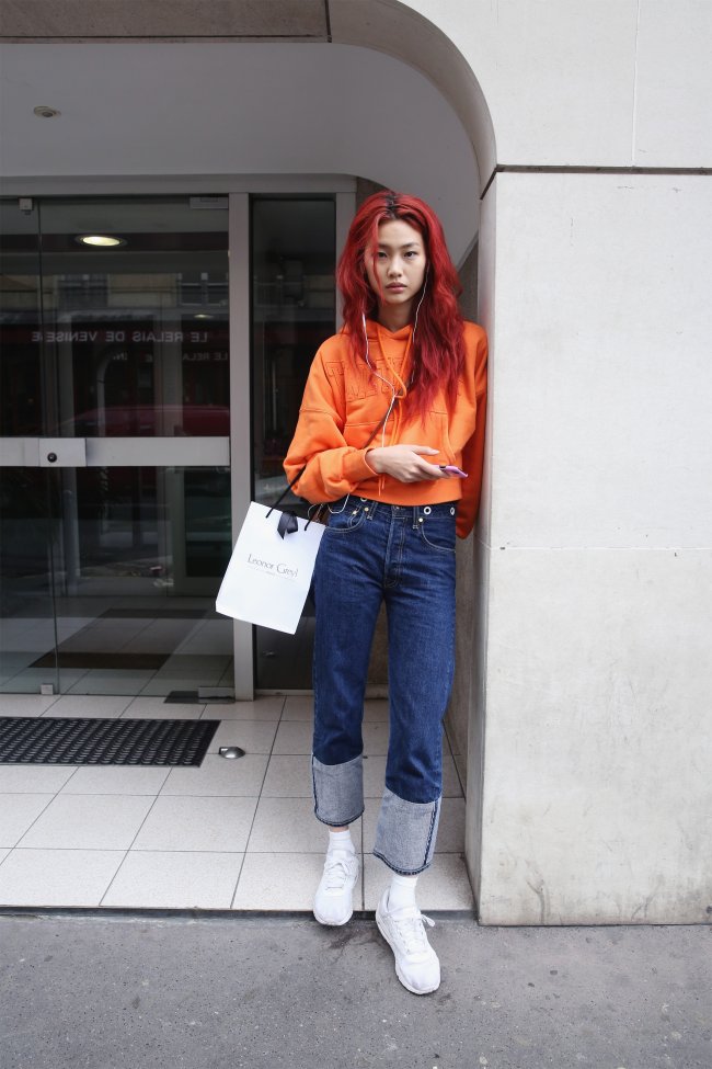 Jung Ho-yeon encostada na parede usando blusa laranja, calça jeans e com uma expressão séria.