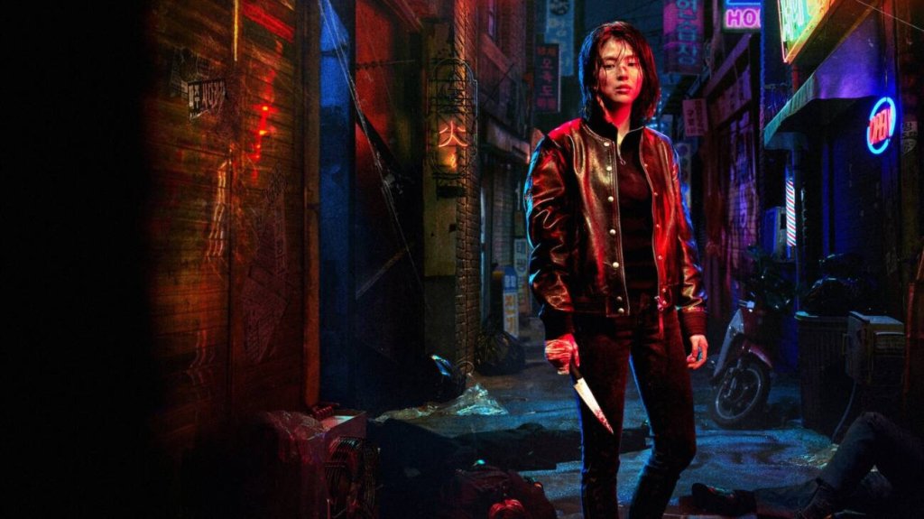 Imagem de divulgação de My Name em que a protagonista está em uma rua vazia de noite segurando uma faca com a ponta ensaguentada; o local está iluminado por luzes vermelhas e azuis; a personagem usa uma blusa com capuz preto e calça também preta