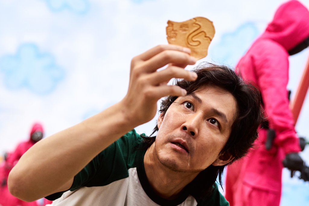 Personagem de Round 6, série sul-coreana da Netflix segurando e observando algo em sua mão; o céu azul claro com nuvens brancas e pessoas com macacão vermelho se destacam ao fundo