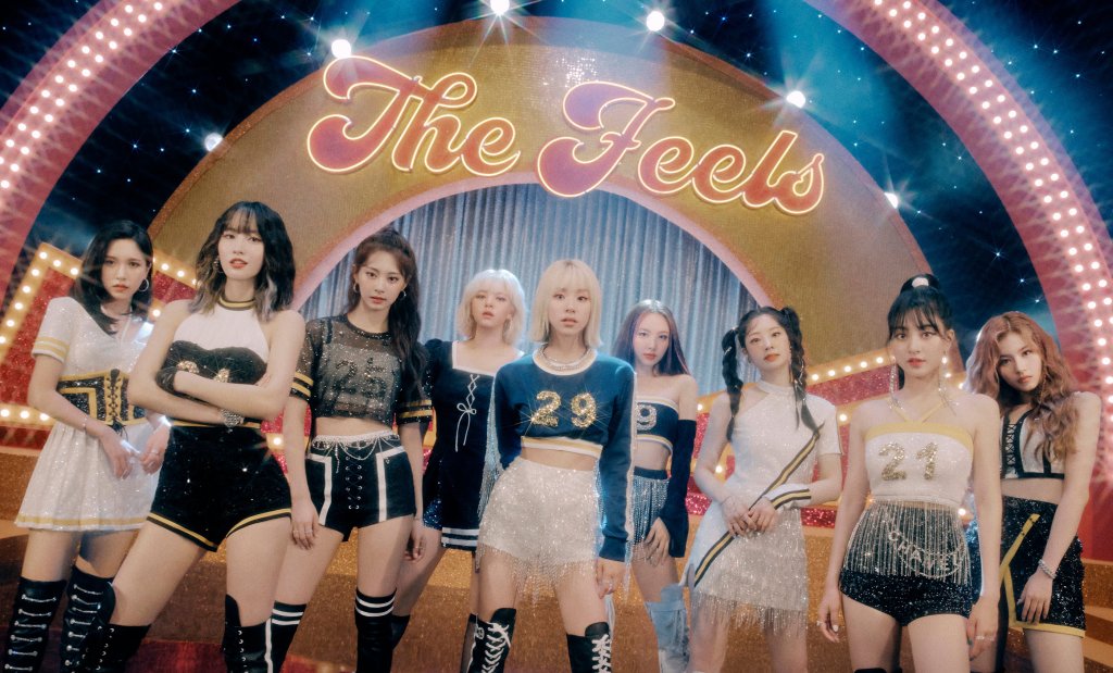 Integrantes do TWICE no clipe de The Feels; elas estão posando na frente de um letreito colorido em vermelho com bordas brilhantes em amarelo; ao fundo é possível ver um círculo amarelo com fitas prateadas na vertical; as cantoras estão olhando para câmera com expressões sérias