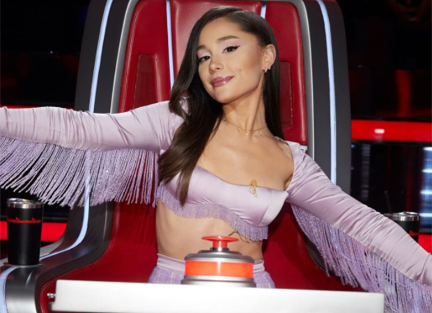 Ariana Grande sentada na cadeira de jurado do The Voice. Ela usa top roxo com franja nas mangas e está com os braços abertos e um leve sorriso no rosto