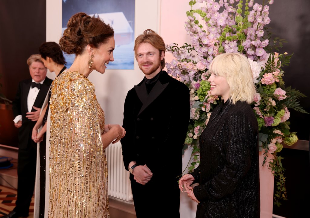 Foto da cantora Billie Eilish conversando com Kate Middleton. Nela, a cantora aparece com uma roupa preta e cabelos loiros conversando com a princesa que está com um vestido dourado e cabelo preso em penteado. Ao fundo, temos uma parede de flores roxas e plantas verdes.