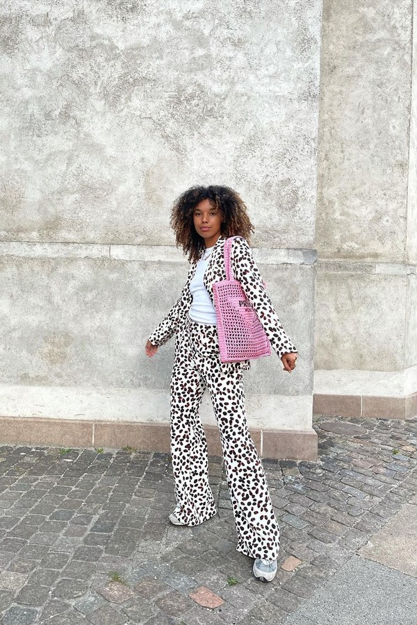 bolsa-de-croche-da-prada Foto de uma mulher em uma calçada. Ela usa o cabelo solto, camiseta branca, casaco branco com estampa de bolinhas pretas, calça com a mesma estampa, tênis e bolsa rosa de crochê da Prada. Ela olha para a câmera e não sorri.