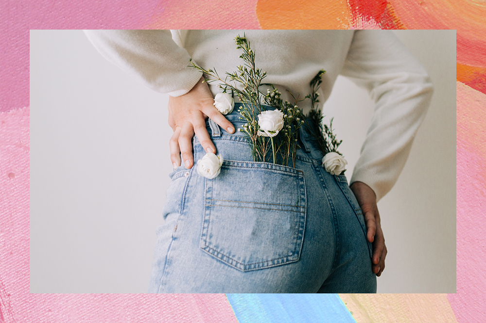 Mulher virada de costas, usando calça jeans com flores brancas dentro do bolso e as mãos no quadril. O fundo da montagem é rosa, laranja e azul