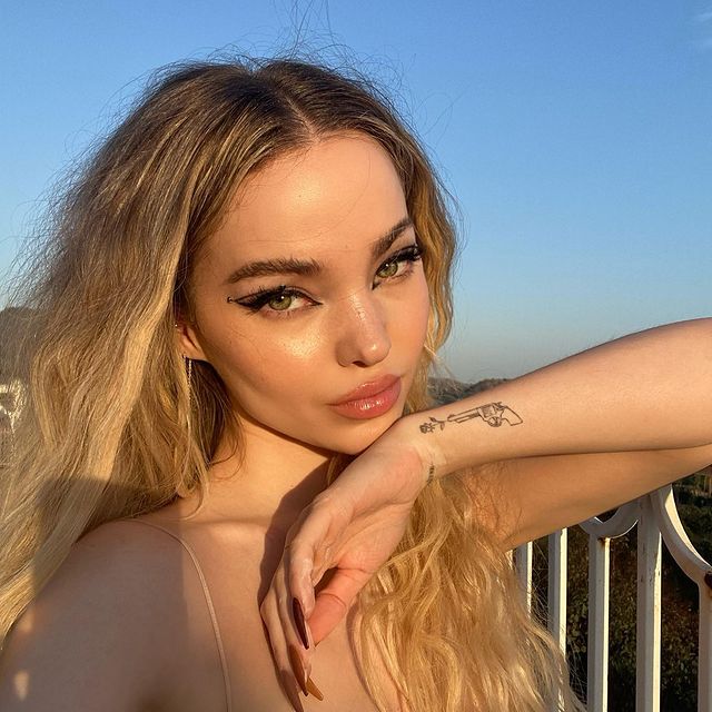 Selfie da Dove Cameron. Ela usa uma blusa de alcinha, cabelo solto e maquiagem com delineado gráfico preto. Ela está com o queixo apoiado em sua mão esquerda, olha para a câmera e não sorri.