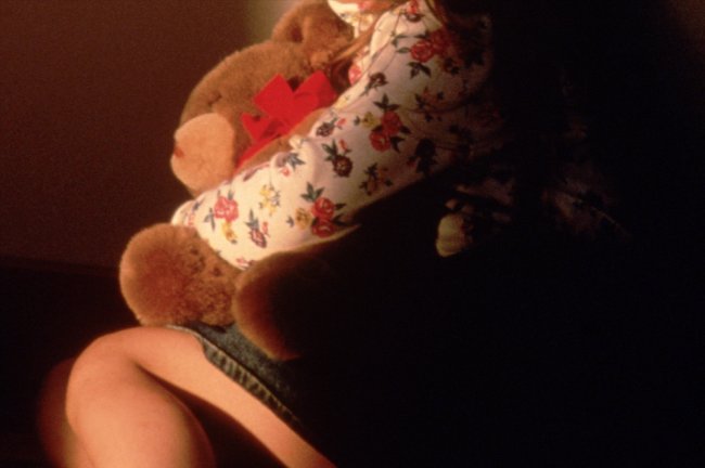 Foto do tronco de uma criança segurando um ursinho de pelúcia. Ela está acuada no canto da sala.