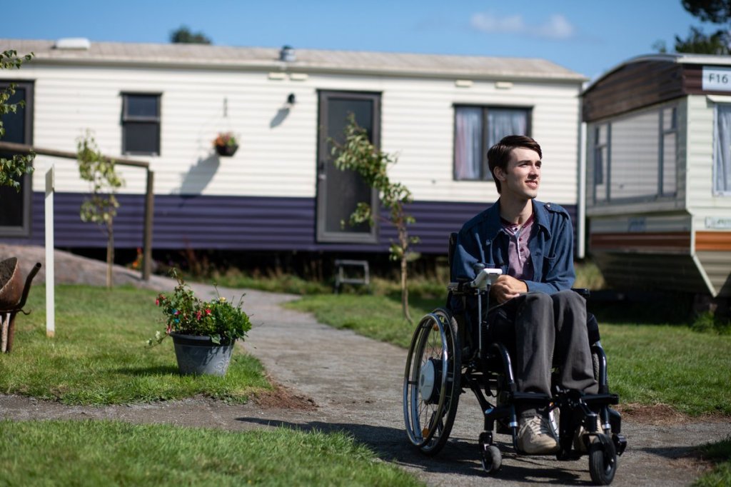 Foto do Isaac, personagem de Sex Education, em frente ao seu trailer. Ele olha para o lado e está sentado em sua cadeira de rodas