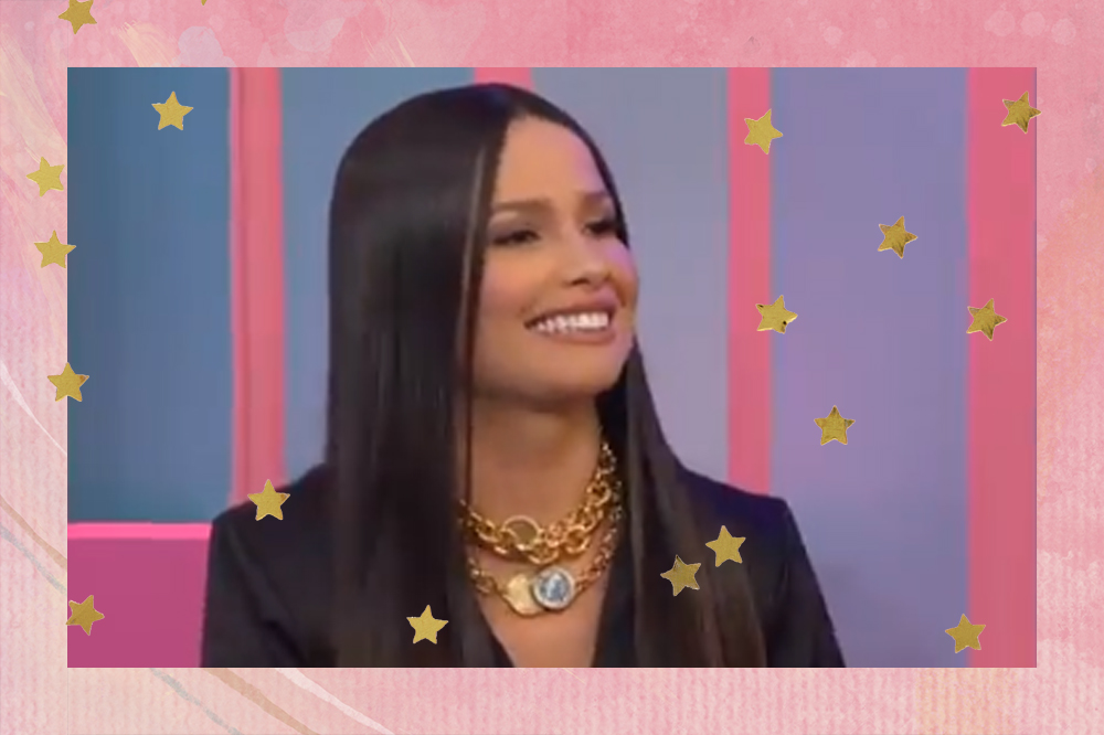 Juliette sorrindo e olhando para o lado no #TVZJuliette, programa do Multishow; ela está com o cabelo solto, usando uma roupa preta com colares dourados; a margem é uma textura em tons de rosa com estrelas amarelas como decoração