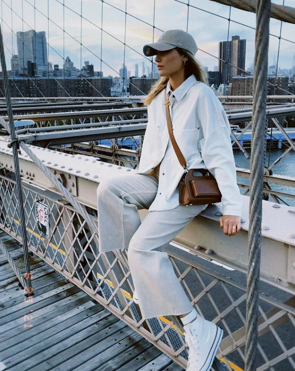 Sasha Meneghel sentada na Brooklyn Bridge, em Nova York, usando boné cinza, camisa off white, calça cinza e tênis branco e uma bolsa marrom atravessada