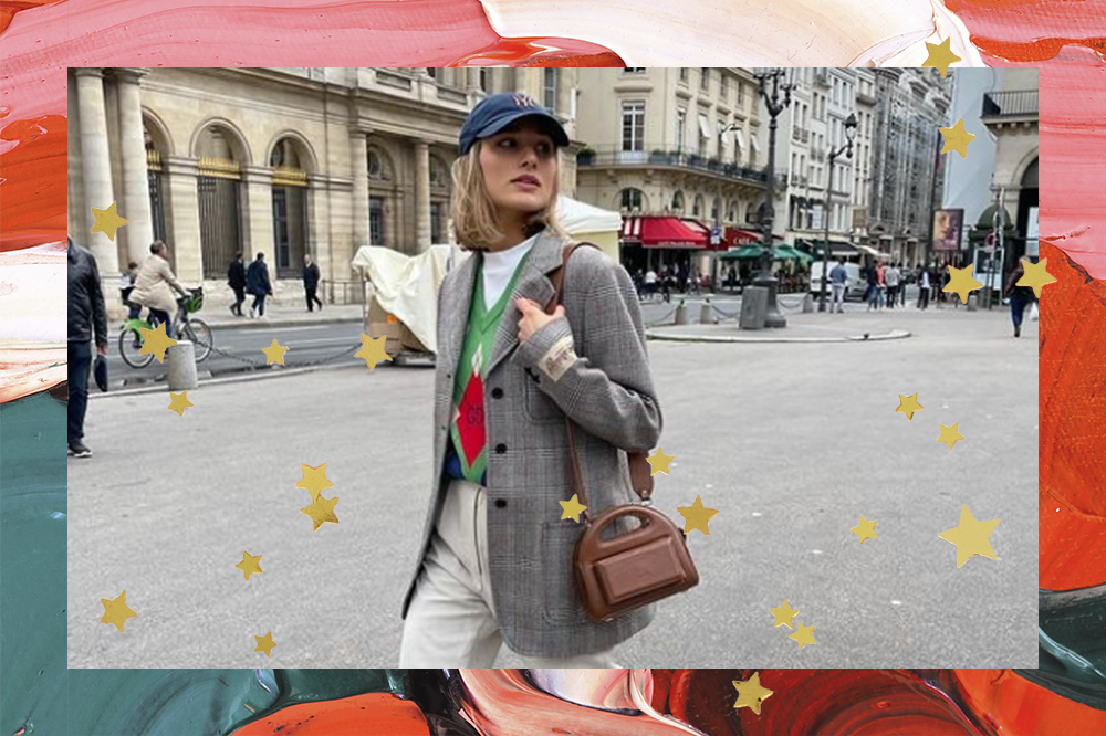 Sasha Meneghel usando blazer xadrez, boné azul e bolsa marrom pendurada em um dos ombros, enquanto olha para o lado na rua. O fundo da montagem é verde, rosa e vermelho e possui estrelinhas douradas