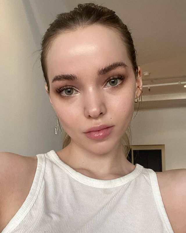 Selfie da Dove Cameron. Ela usa uma blusa branca, cabelo preso e maquiagem com efeito natural. Ela olha para a câmera e não sorri.