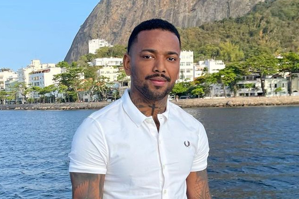 Imagem de Nego do Borel sorrindo levemente; ele está usando uma camisa branca e o fundo da imagem mostra o mar e parte do morro do Pão de Açúcar no Rio de Janeiro