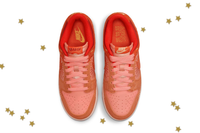 Foto com fundo branco e com estrelinhas douradas. No centro, temos o novo tênis da Nike do modelo Dunk Low com as cores laranja, vermelho e detalhes em amarelo.