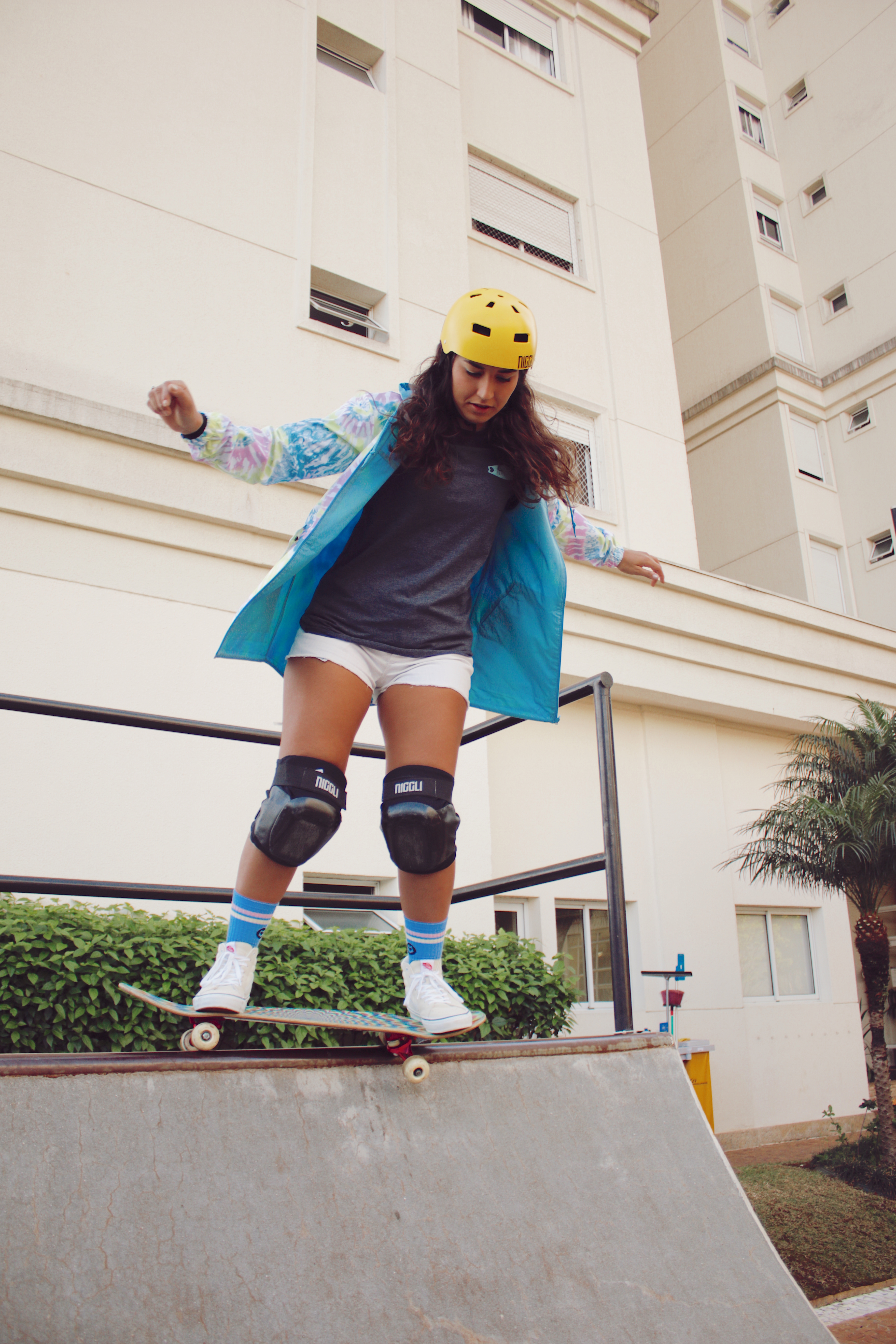 Thais Gazarra andando de skate em uma mini rampa com jaqueta azul, camiseta cinza, short e capacete e joelheira