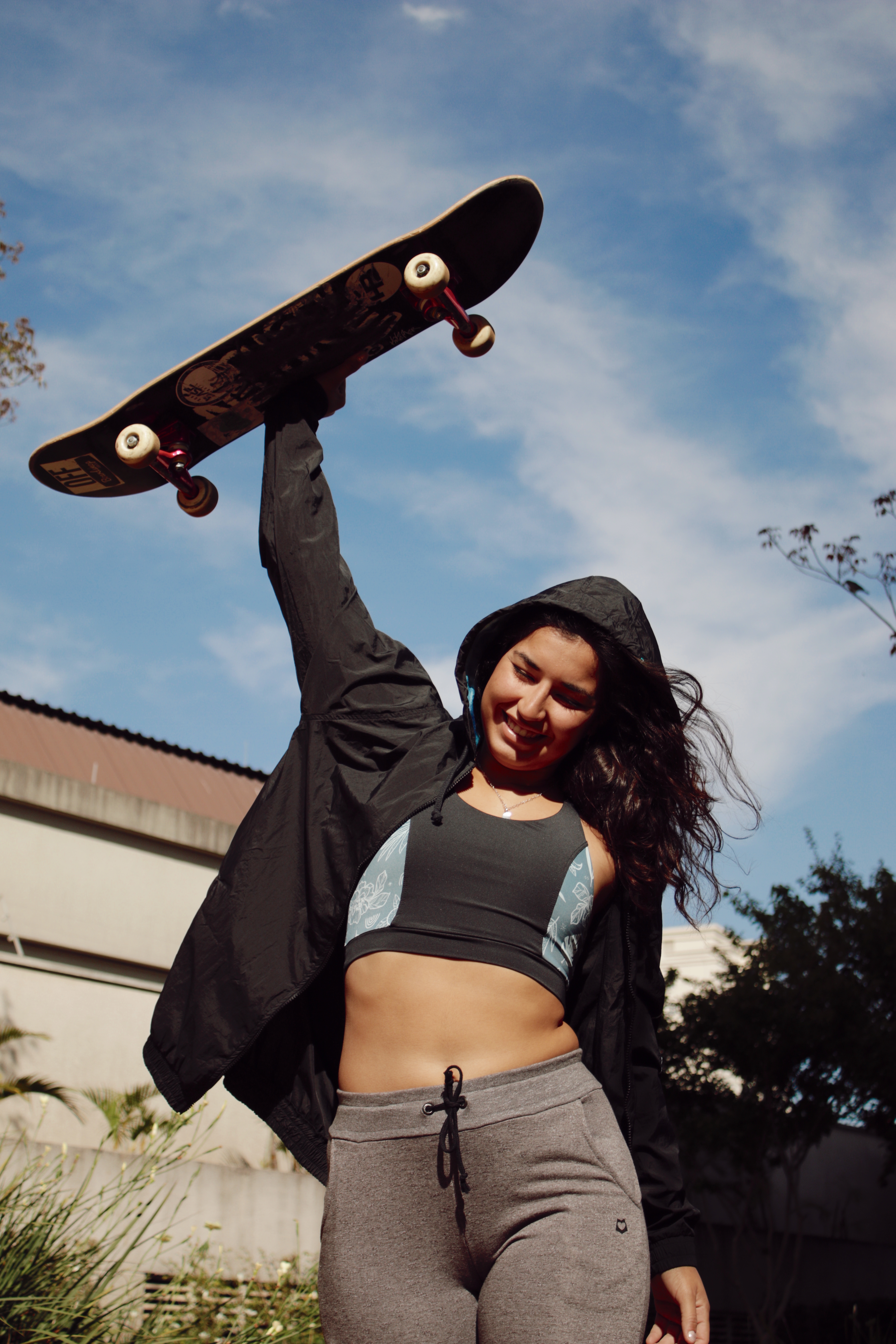 Thais Gazarra sorrindo, usando um top e uma calça cinza e uma jaqueta preta enquanto segura um skate com uma das mãos e o levanta pra cima