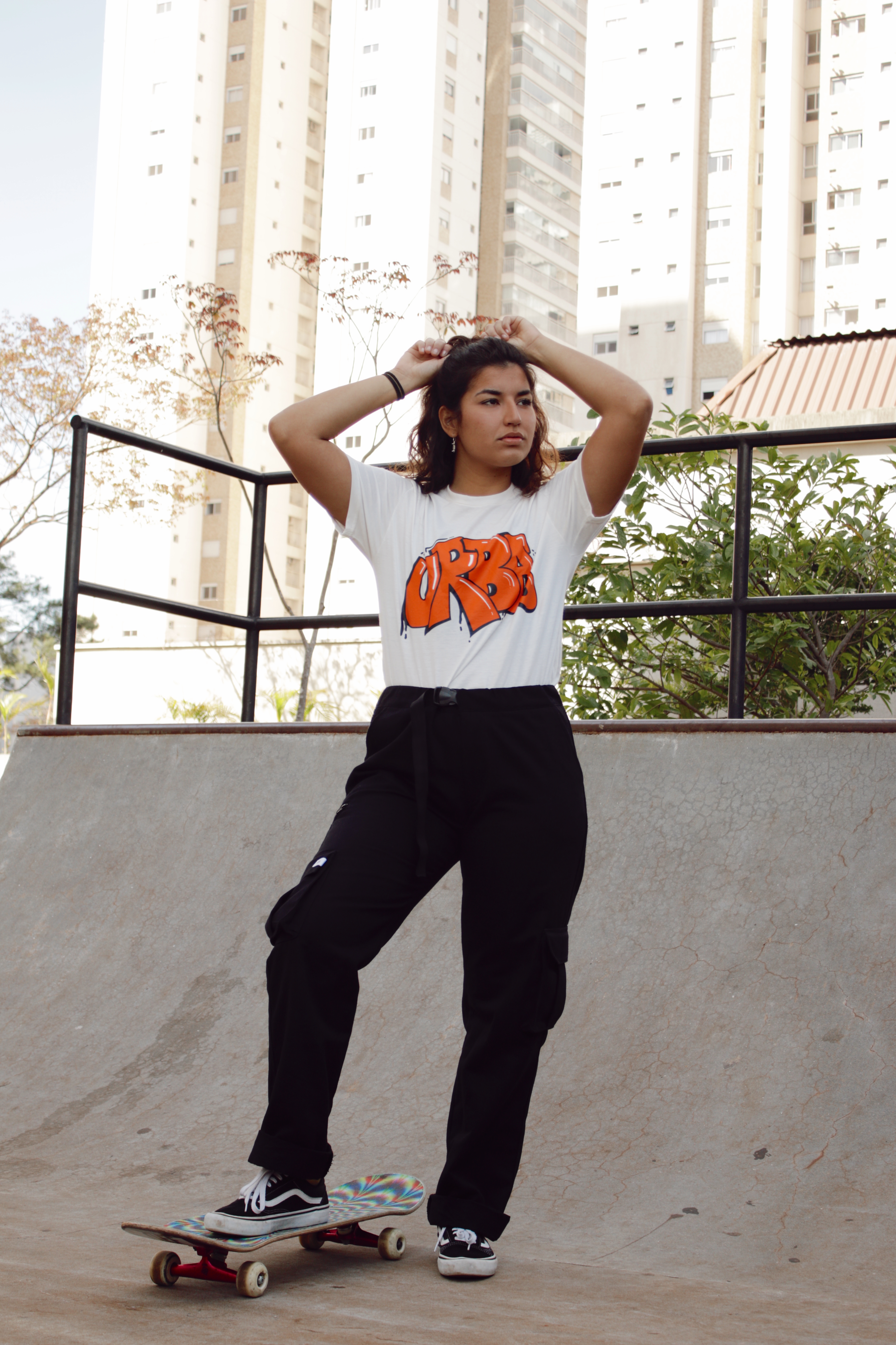 thais-gazarra-skate-58 Thais Gazarra em mini rampa de skate usando camiseta branca, calça cargo preta, com um dos pés apoiado no skate e mexendo no cabelo com as duas mãos para trás