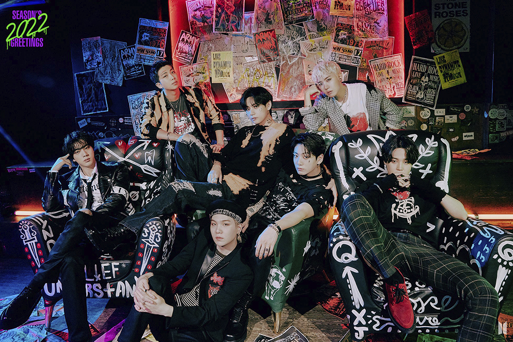 Foto de divulgação do grupo BTS para o AMAs 2021; eles estão usando roupas pretas, expressões sérias e posando sentados em um fundo com folhas de jornais colados na parede; uma iluminação vermelha e azul estão na parte superior da imagem
