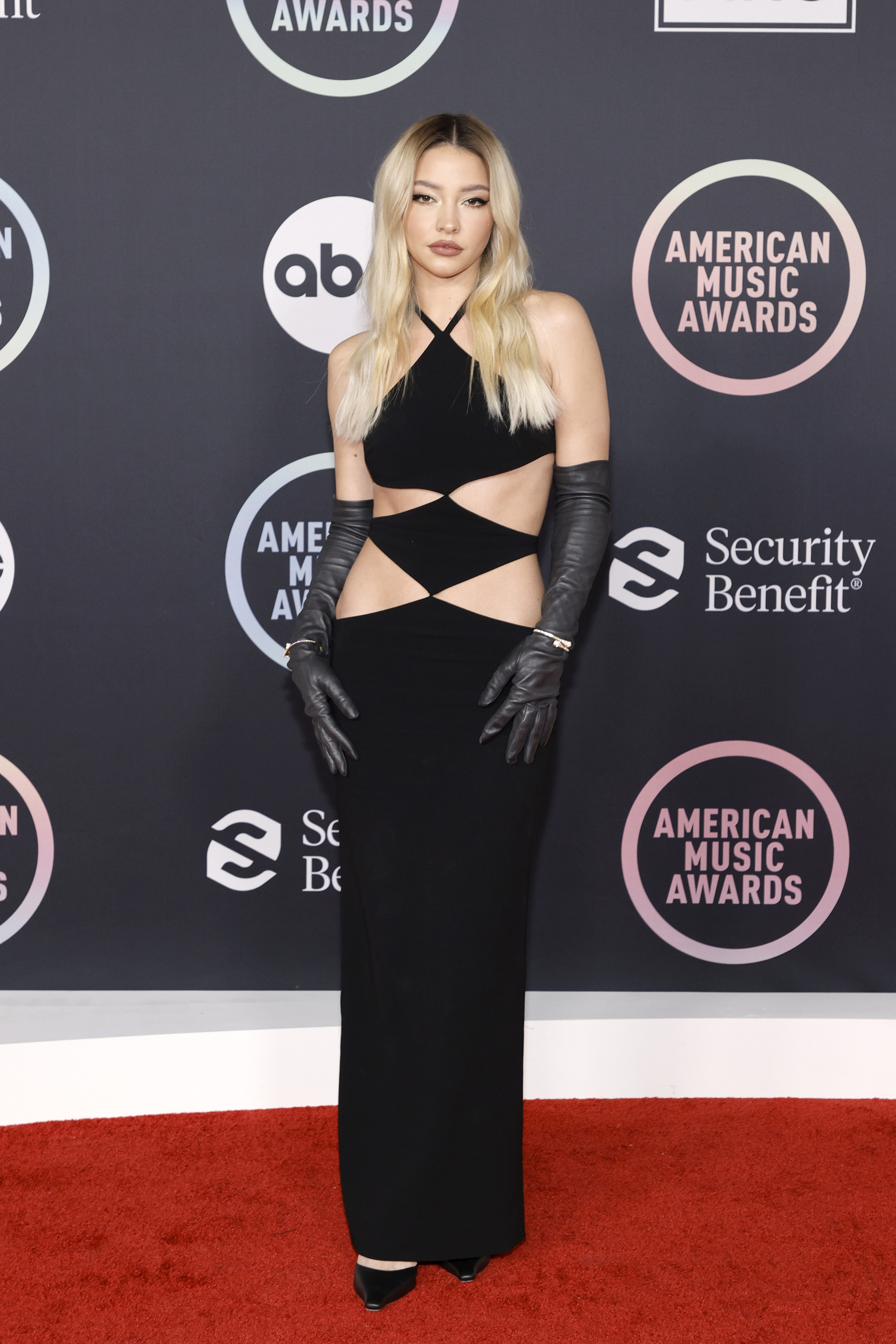 Madelyn Cline no American Music Awards 2021 usando vestido preto com recortes na barriga e luvas pretas até o cotovelo