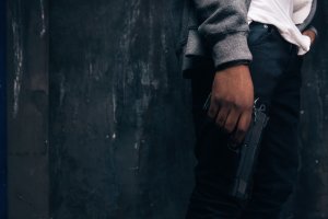 Foto da mão de um homem segurando uma arma de fogo. Dá pra ver que ele usa um moletom cinza.
