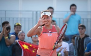 Foto da tenista chinesa Peng Shuai. Ela está na quadra, veste um conjunto de blusa e short salmão, segura a raquete com a mão esquerda e acena para a câmera com a direita. Ela está sorrindo.
