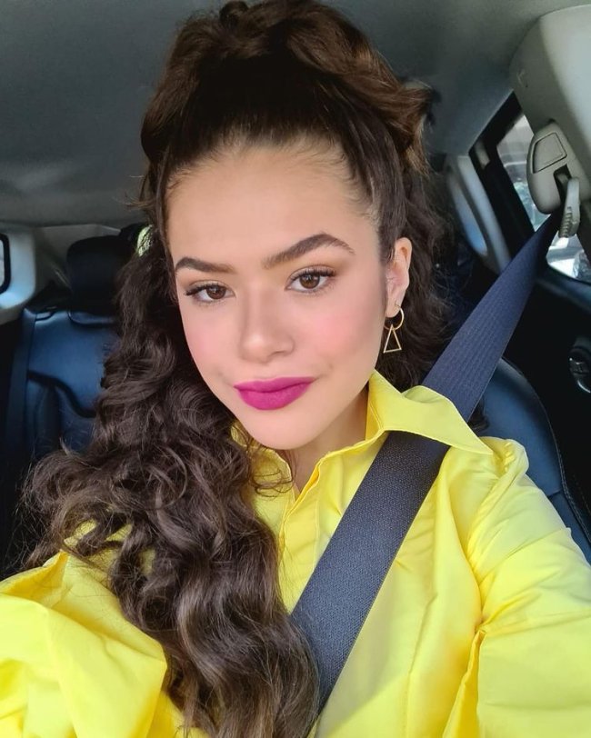 Maisa com penteado meio preso Maisa posando para selfie dentro do carro, podemos ver o cinto de segurança, e ela usando blusa amarela com penteado meio preso e expressão sorridente.