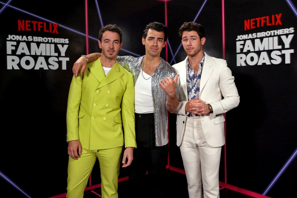 Os irmãos aparecem posando para foto. Kevin à esquerda com terno verde, Joe no centro com camisa branca e jaqueta prateada e Nick à direita com terno branco e camisa estampada.
