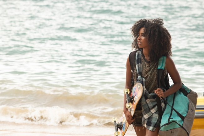 Imagem de cena com Gabz como Yasmin em Temporada de Verão, nova série da Netflix; ela está andando na praia segurando um skate; uma bolsa cinza com azul/verde; ela usa um shorts jeans com uma blusa estampada e uma blusa xadrez preto e branco amarrada no corpo; ao fundo é possível ver o mar e areia