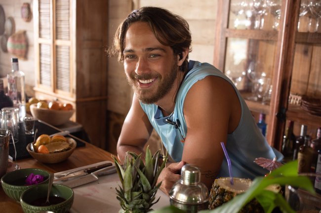 Imagem de cena com André Luiz Frambach como Miguel em Temporada de Verão, nova série da Netflix; ele está sorrindo para cãmera e usa ele apoiado em um balcão ou uma mesa com algumas plantas;