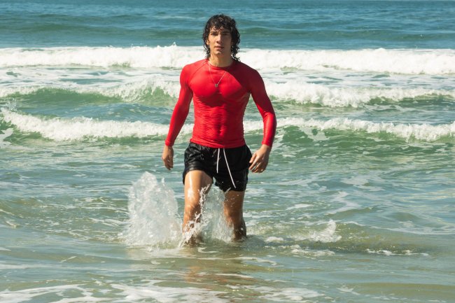 Imagem de divulgação de cena com Jorge Lopez como Diego em Temporada de Verão, nova série da Netflix; ele está saindo do mar com a roupa molhada e veste uma bermuda preta com uma camisa vermelha de manga comprida; ao fundo é possível ver o mar e a espuma de algumas ondas