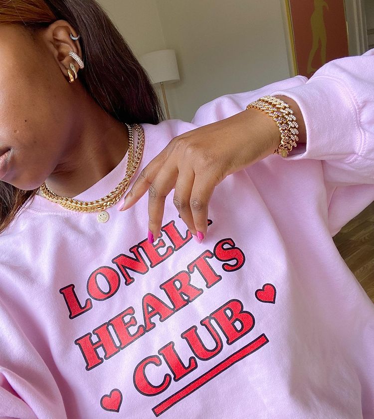 Garota usando colares e pulseiras de correntes e um moletom rosa escrito Lonely Hearts Club em vermelho