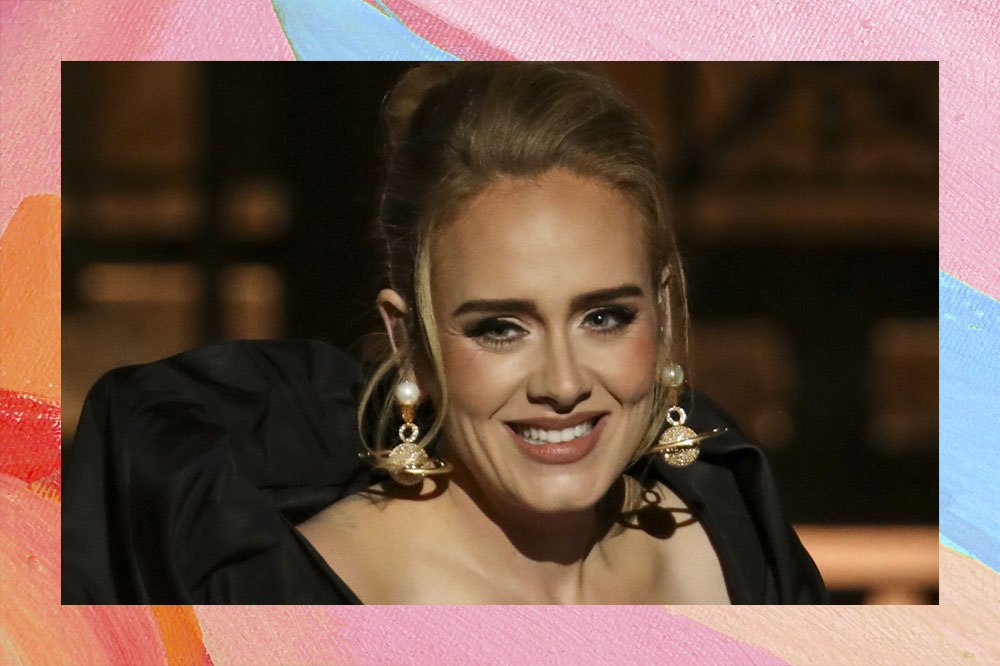 Foto de Adele com cabelo preso e liro, brincos grandes e roupa preta. Ela está sorrindo.
