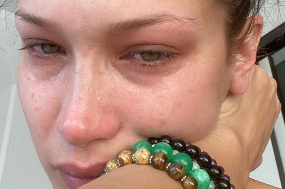 Foto de Bella Hadid chorando, com os olhos inchados, cabelo preso em coque e pulseiras verdes, pretas e marrons.