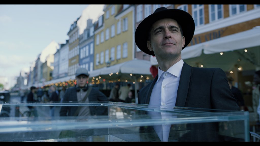 Berlim, de La Casa de Papel em uma cena da série; ele está sorrindo levemente usando uma camisa branca e um chapéu e paletó na cor preta; ele está olhando para o horizonte em pé em um barco branco
