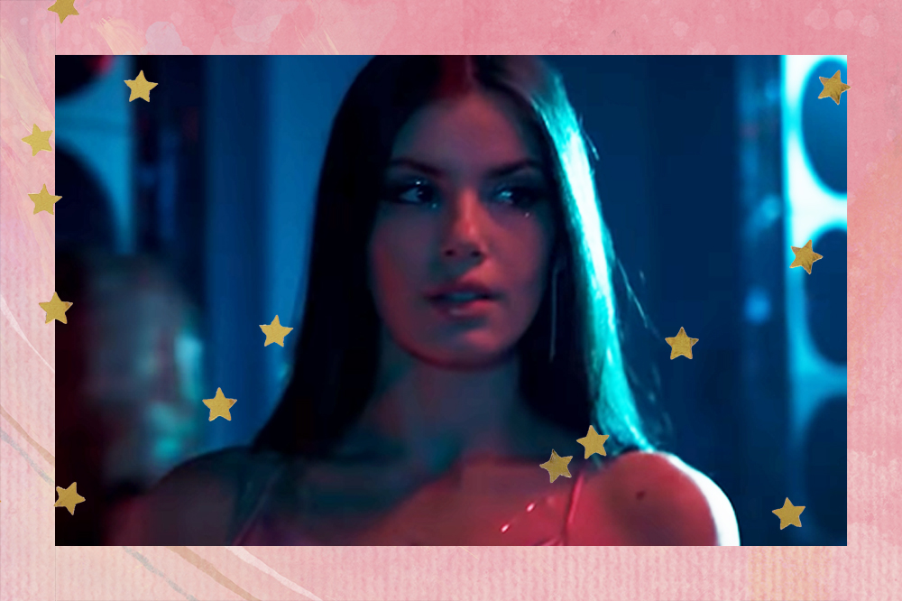 Camila Queiroz como Angel em Verdades Secretas 2; ela está em um ambiente fechado com luzes azuis, parecendo uma balada ou um evento; com o cabelo solto dividido ao meio e usando uma maquiagem brilhante com vestido de alças; a margem é uma textura em tons de rosa e estrelas amarelas decoram a imagem