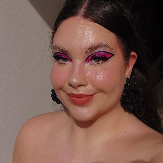 Selfie de uma mulher. Ela está com o cabelo preso, usa um brinco de argola preto e usa uma maquiagem básica com delineado preto banana e sombra rosa.