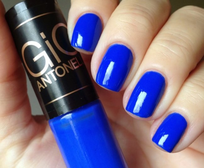 Foto de uma mão segurando um esmalte azul da marca Gio Antonelli com as unhas da mesma cor.
